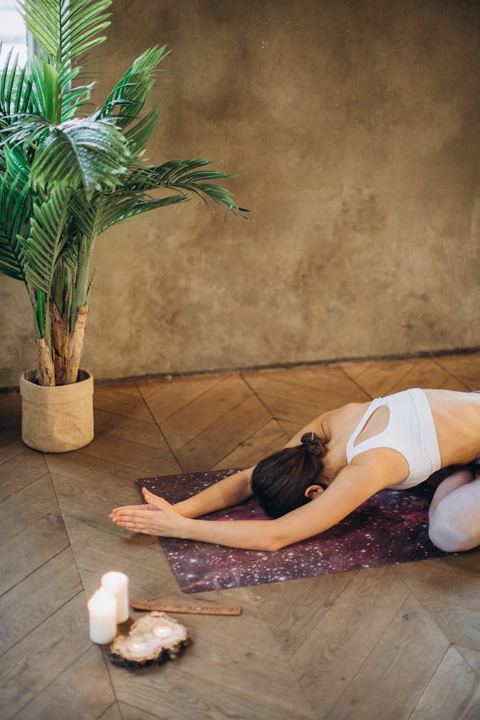 Woman in child's pose on yoga mat with candles and plant, creating a serene indoor setting.